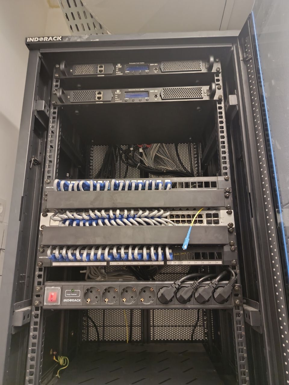 24U basement rack at Ants Pants during commissioning, January 2026 — amplifiers at the top, laced patch panels, the 48-port PoE switch with the yellow optical uplink to the Family Nest core, and a surge-protected PDU below