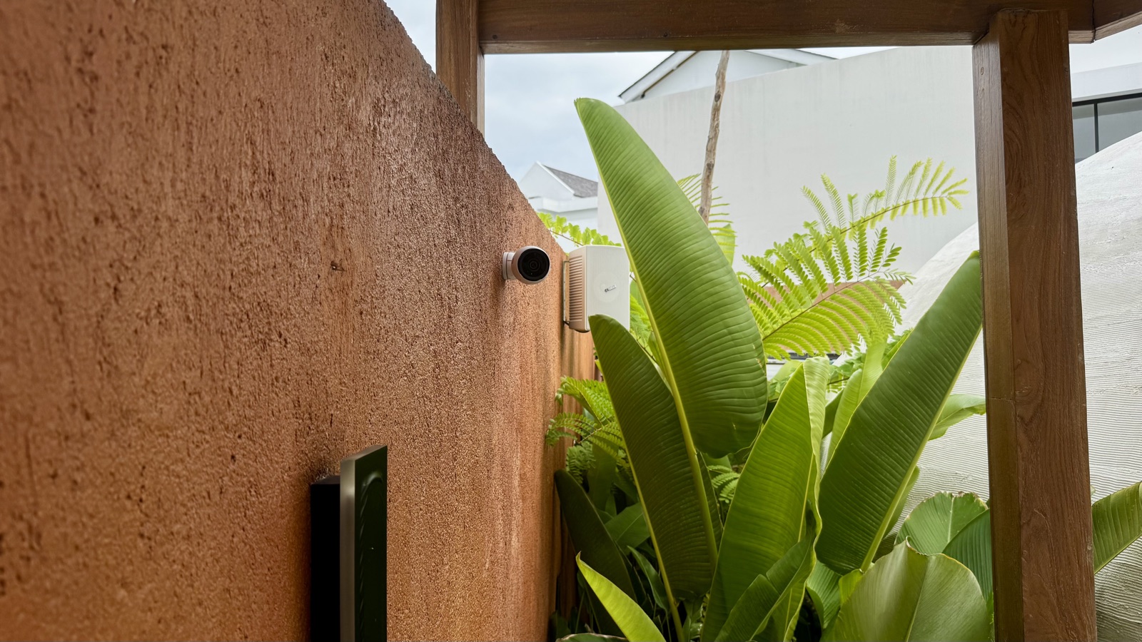 UniFi bullet camera and a white loudspeaker sharing a garden wall at ATMOS, a steam dome behind the foliage — surveillance and estate audio on the same low-voltage backbone