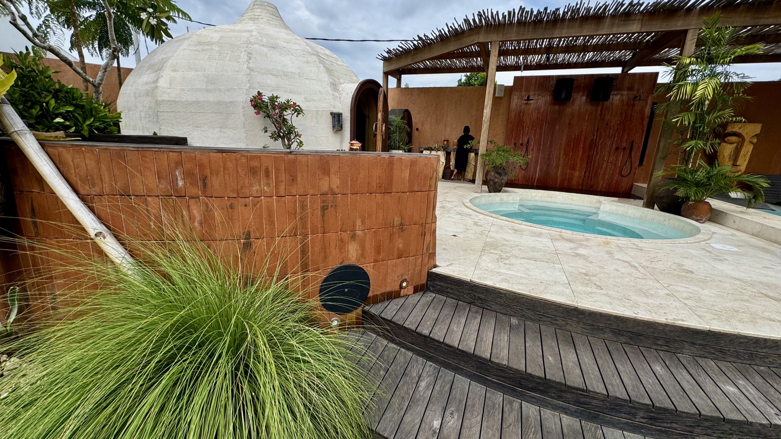 White steam dome and plunge pool at ATMOS Social Steam Club by day — terracotta brick wall with a recessed round speaker grille at the deck edge, rain showers under the pergola behind