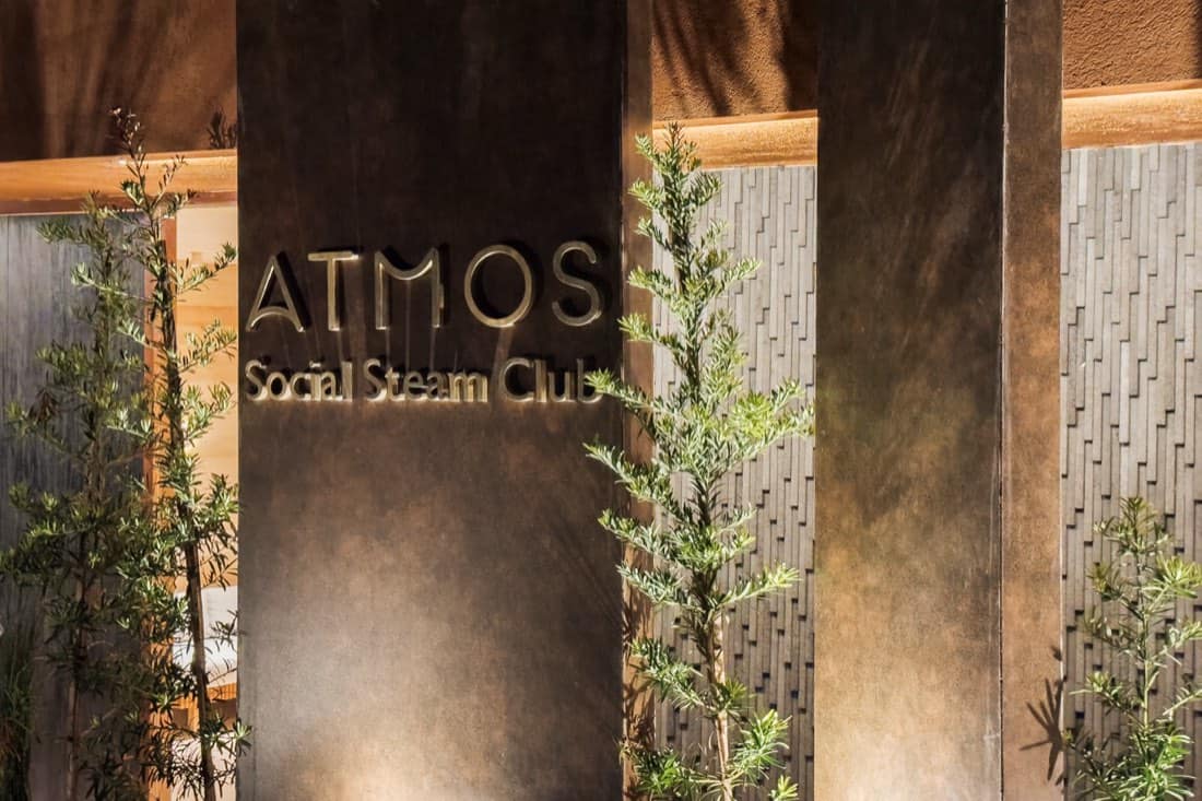 Close-up of the illuminated brushed-gold ATMOS Social Steam Club sign mounted on a patinated metal panel beside a textured tile wall and greenery