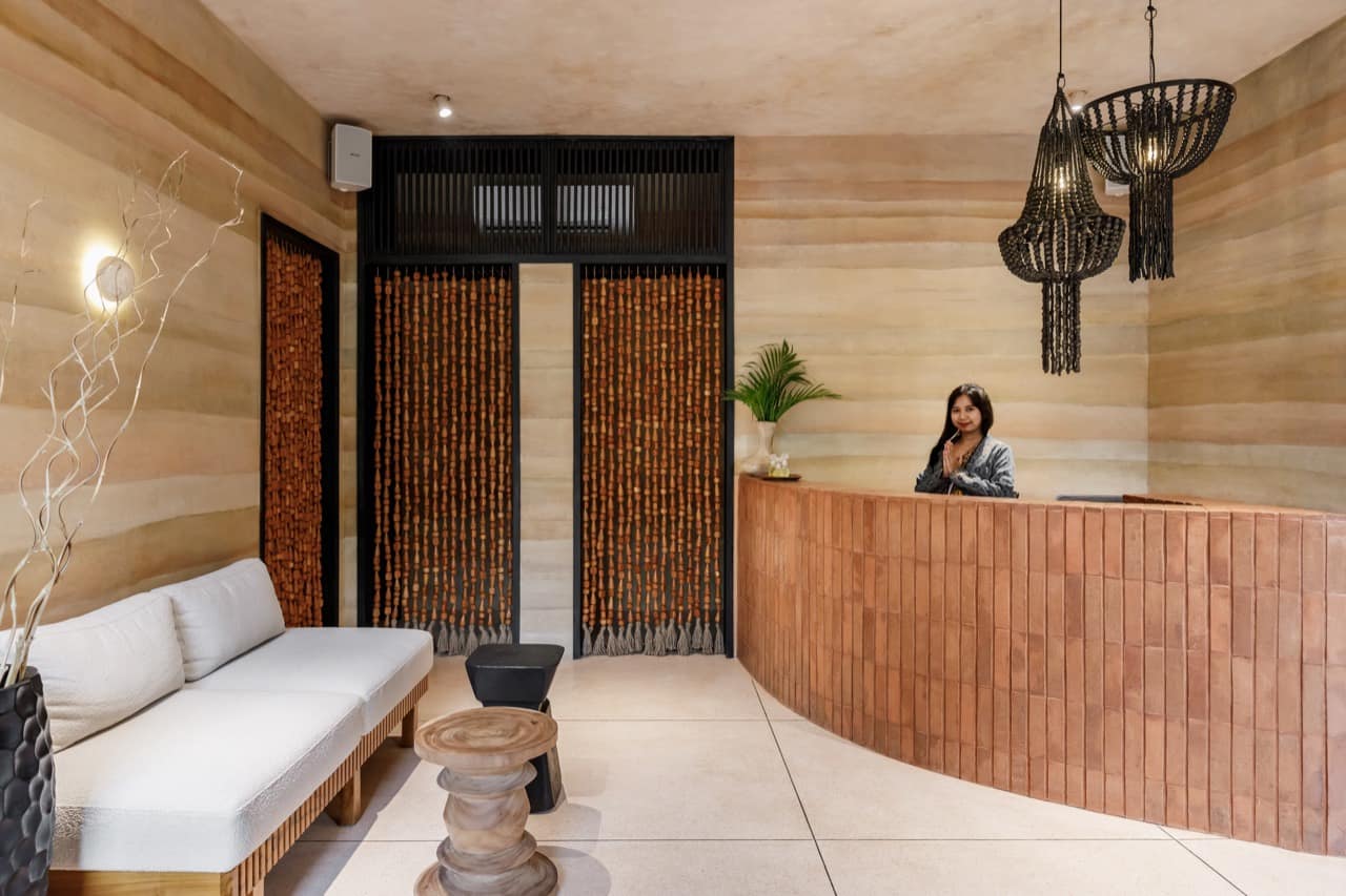 Reception lounge at the ATMOS Social Steam Club with a curved terracotta brick desk, beaded chandeliers, rammed-earth walls, a white upholstered bench and carved wood stools, and a staff member greeting guests