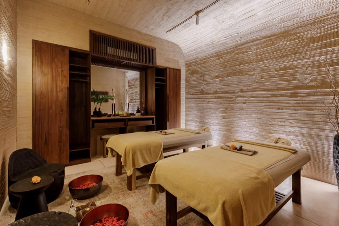 Couples treatment room at the ATMOS Social Steam Club with two massage tables draped in mustard towels, textured rammed-earth walls and ceiling, a wooden vanity with mirror, and brass bowls of red flower petals on the floor