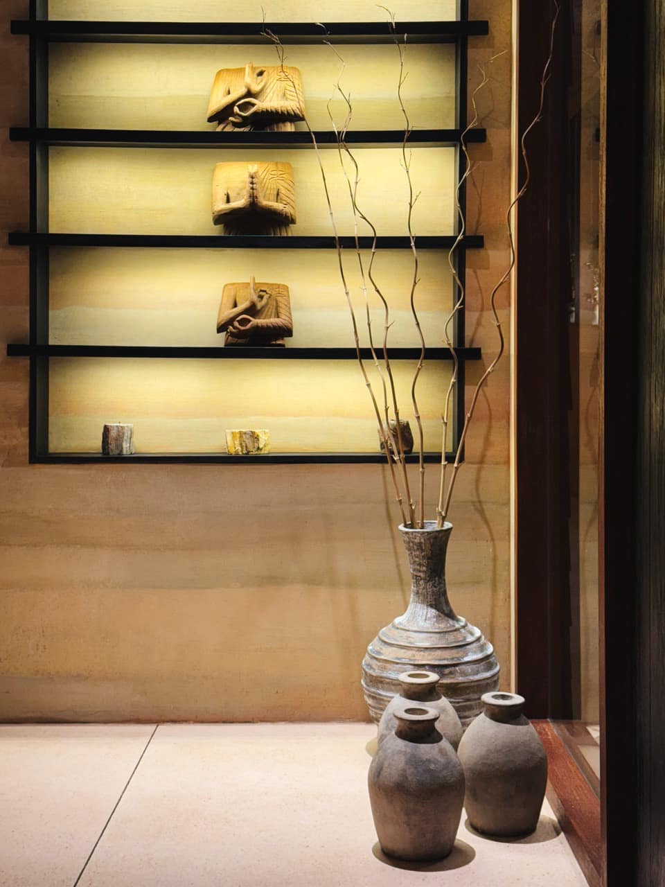 Decorative niche at the ATMOS Social Steam Club with backlit shelves displaying carved wooden figures, beside aged stone pots and a tall vase of bare branches against a rammed-earth wall