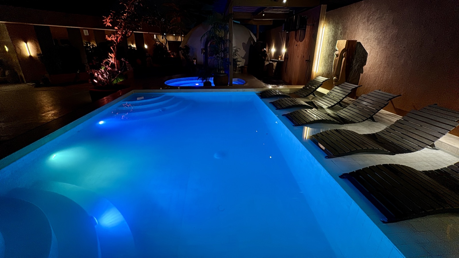 ATMOS pool at night lit deep blue by the RGB scene, black loungers along the edge and warm linear wall lights behind — the plunge pool glowing violet in the background