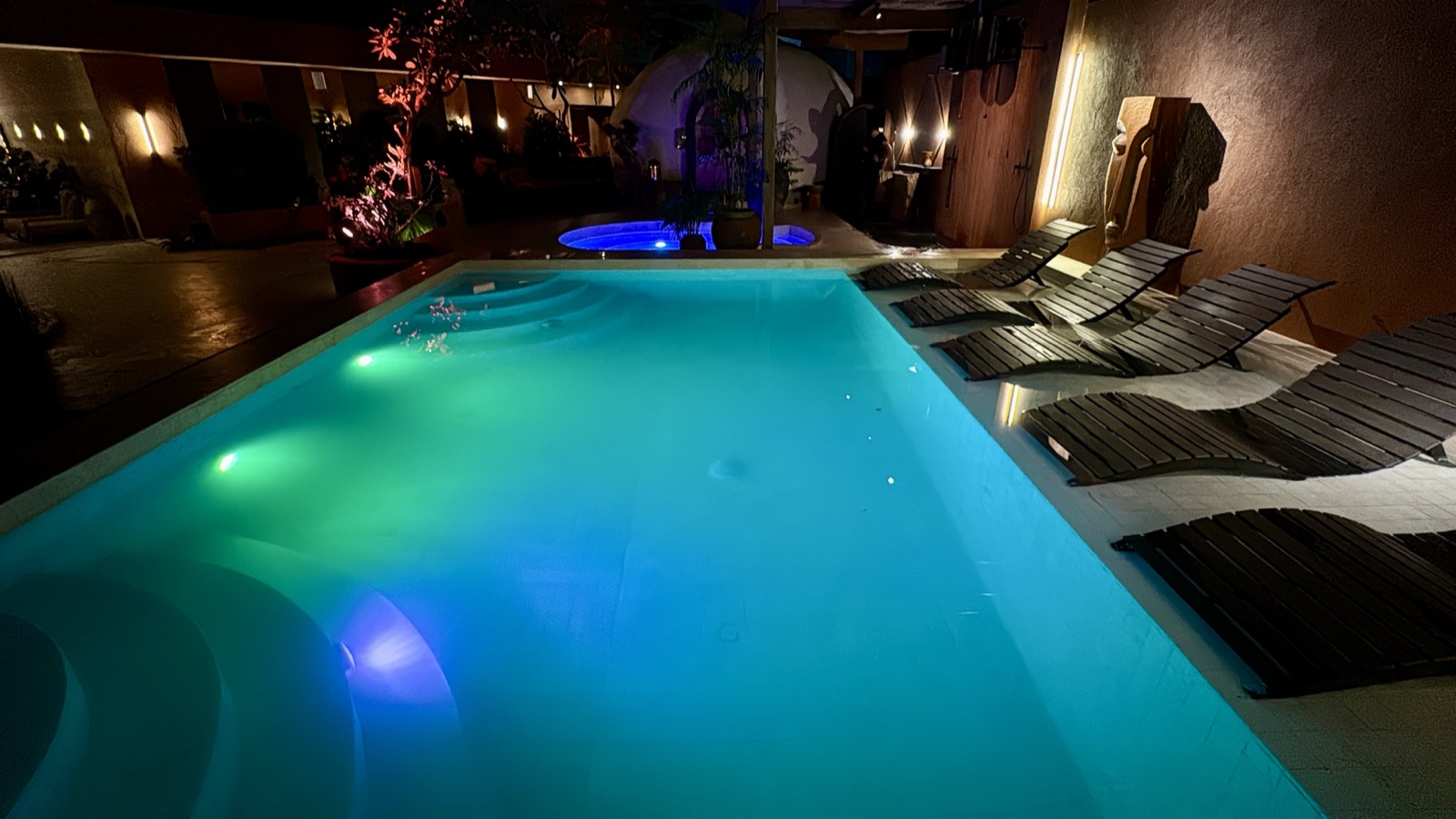 The same ATMOS pool in a turquoise-green RGB scene minutes later — two colour states of the pool lighting, loungers and rammed-earth walls unchanged