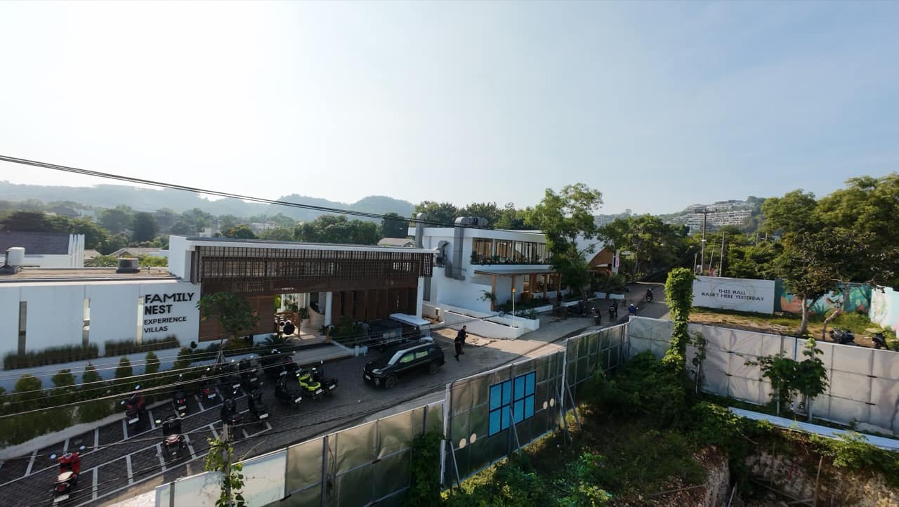 Drone view of the Family Nest Experience Villas frontage, showing the building's white render wall with FAMILY NEST EXPERIENCE VILLAS signage, a slatted-timber facade, motorbike parking, and green hills behind