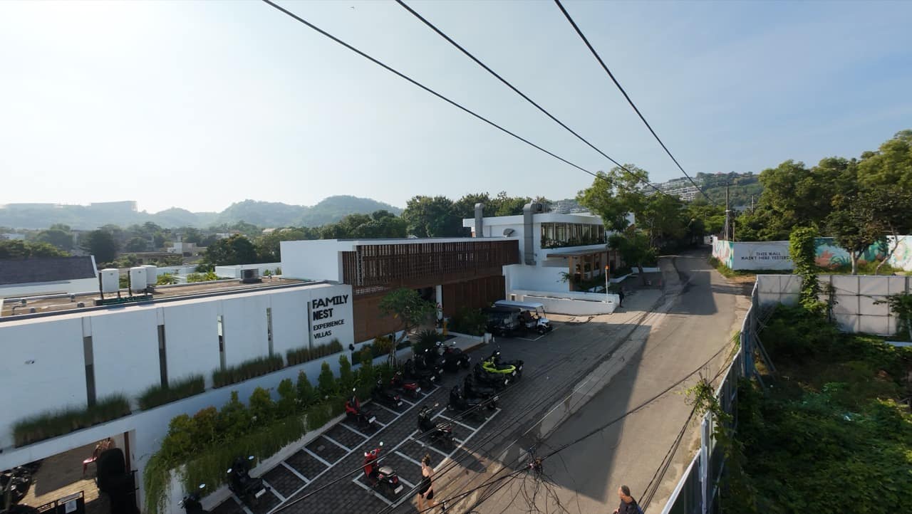 Aerial shot of the Family Nest Experience Villas entrance and parking forecourt with branded signage on the white facade, a row of motorbikes, and the surrounding Bali hillside in the distance