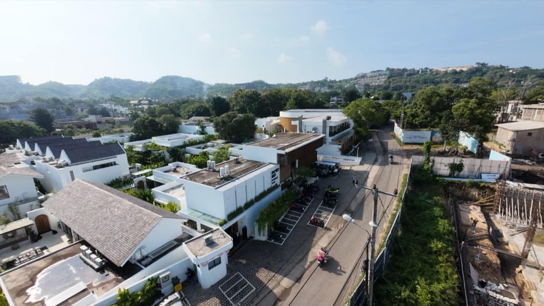 Elevated aerial of the Family Nest resort showing white villas with pitched grey roofs, the timber-roofed clubhouse, a roadside parking row, and forested hills under a clear sky