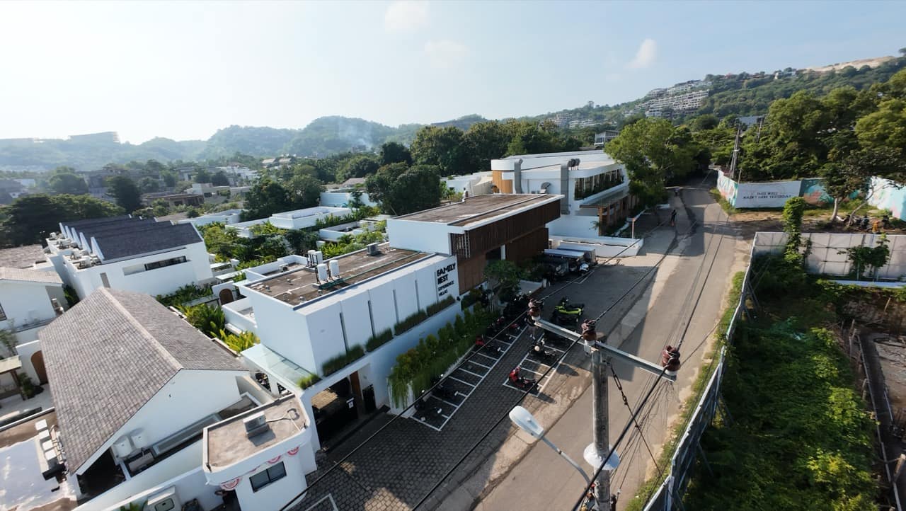 High aerial over the Family Nest resort and neighbouring white villas with rooftop terraces, the curved timber clubhouse, and a winding lane leading toward the green hilltops of Bali
