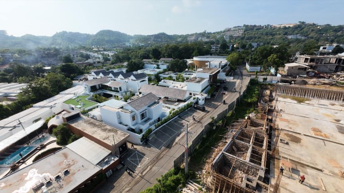 Wide aerial of the Family Nest resort district with rows of white villas and rooftop lawn terraces spread across a green Bali hillside under a dramatic blue and cloud-filled sky