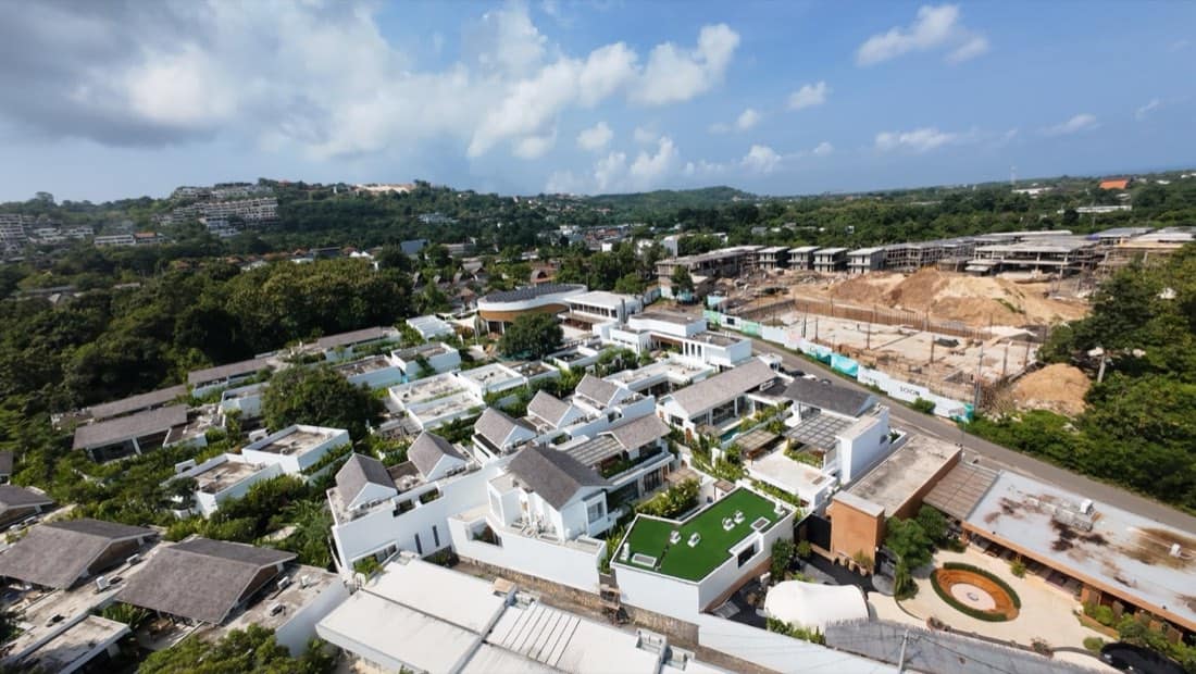 Aerial overview of the Family Nest resort showing the timber clubhouse and white villas with rooftop gardens, bordered by trees and a hillside, with construction land to the right