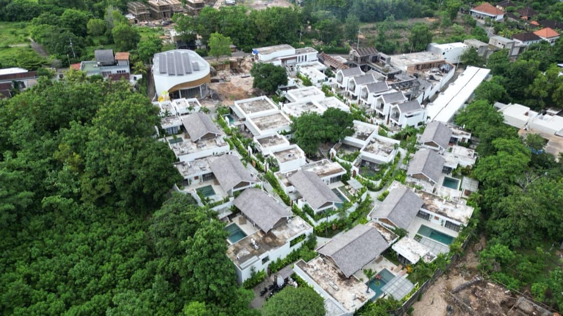 Aerial view of the Family Nest resort villas with grey thatched roofs, private pools and rooftop terraces on a green Bali hillside in Pecatu, Uluwatu