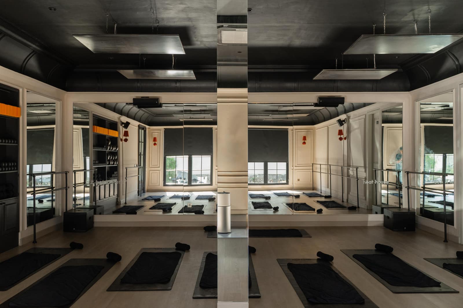 Heat Space studio room in daylight showing the white-panelled walls, windows with roller blinds, ceiling-mounted infrared heating panels and black mats laid out across the timber floor