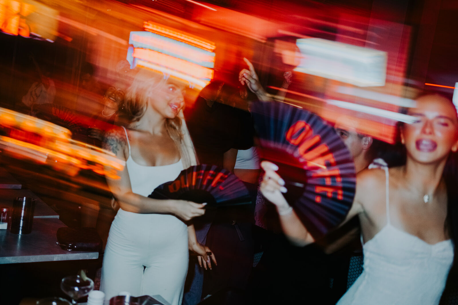 Guests dancing at OUFFF Burger late at night under red neon light, holding branded OUFFF hand fans, with the music at the centre of the room