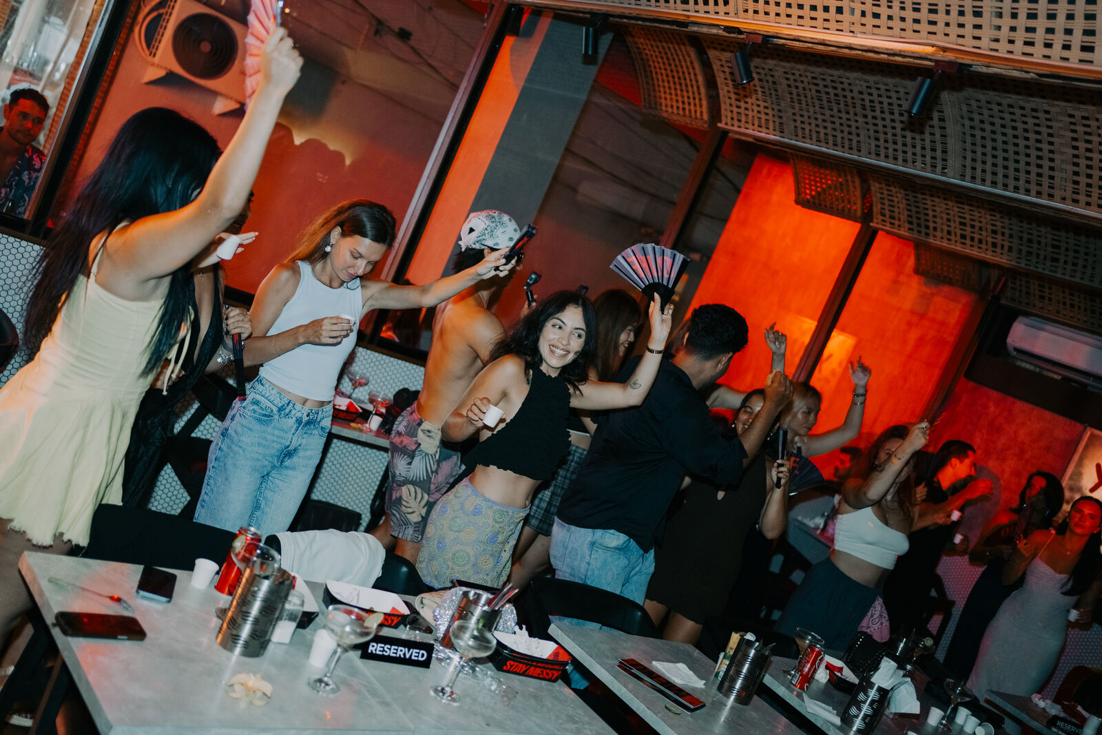 A packed OUFFF Burger at night — guests dancing between marble-top tables under warm and red light, with woven rattan and perforated ceiling panels overhead and no visible speakers