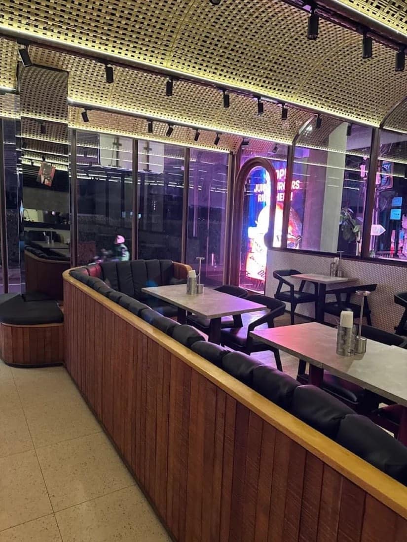 Interior of a modern burger venue at night with a curved woven rattan ceiling, warm strip lighting, dark leather banquette seating, wooden paneling, stone-top tables and a glass wall showing purple neon and street signage outside