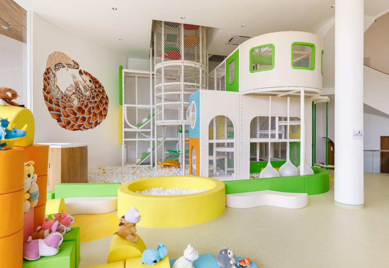 Main indoor play hall of the Pangolin Kids Club with a large painted pangolin mural, a multi-level netted climbing structure, a yellow ball pit, and shelves of soft toys on the green floor