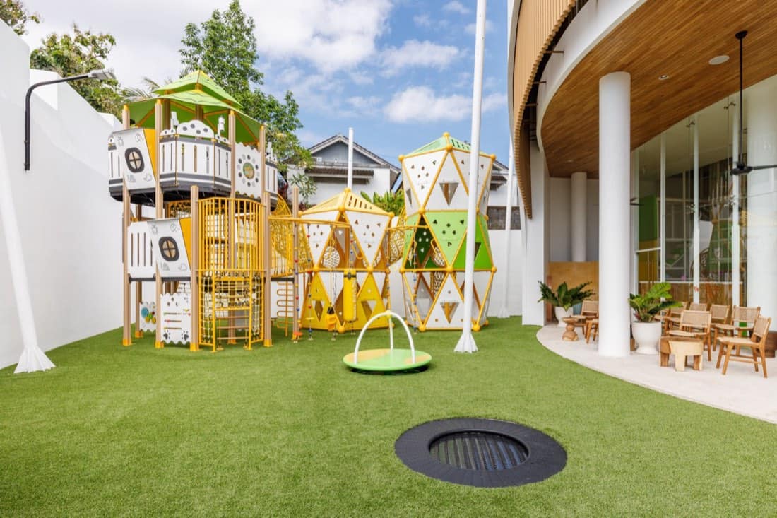 Outdoor playground at the Pangolin Kids Club with yellow-and-white climbing towers and geometric net panels on artificial turf, an in-ground trampoline, and wooden cafe tables under the curved timber building eave