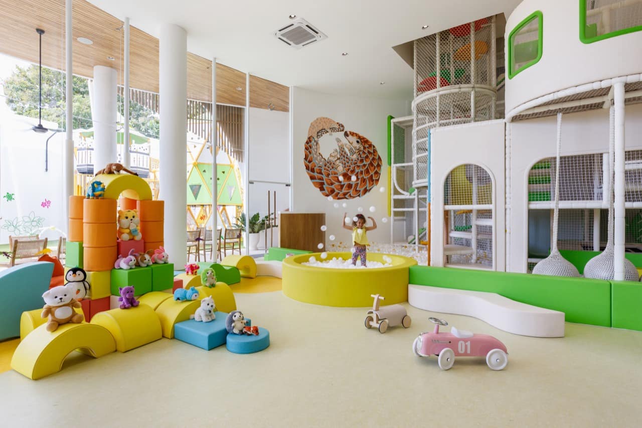 Bright play area inside the Pangolin Kids Club where a child plays in a round white ball pit beneath the pangolin mural, with soft foam blocks, plush toys, ride-on toy cars, and the two-storey play structure