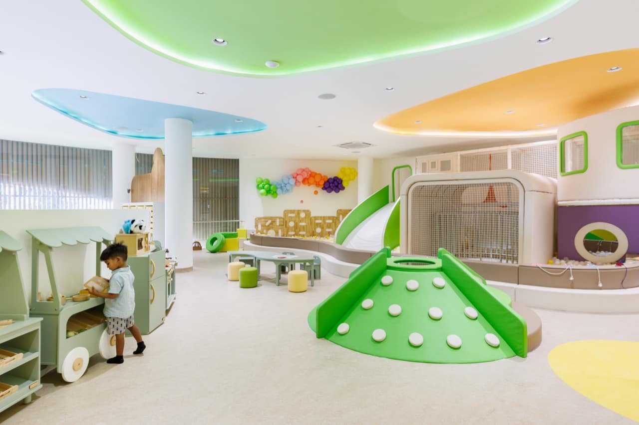 Toddler soft-play zone at the Pangolin Kids Club with a green dome climber, a balloon garland on the wall, a wooden play kitchen, and coloured ceiling discs lit in green, blue and orange, with a young child playing