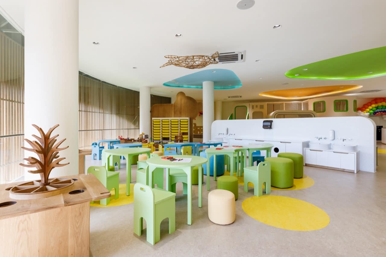 Craft and activity room at the Pangolin Kids Club with rounded green child-sized tables and chairs, a wooden play island, storage cubbies, sinks along the wall, and pastel ceiling discs