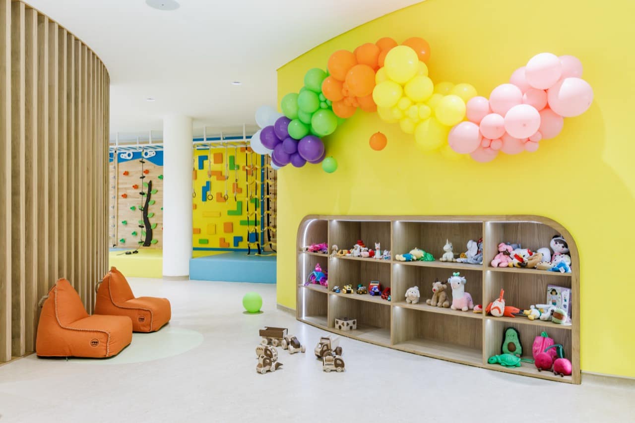 Climbing and toy corner of the Pangolin Kids Club with two yellow climbing walls, a sunny yellow accent wall topped by a pastel balloon garland, orange bean-bag chairs, and a low curved shelf full of plush toys