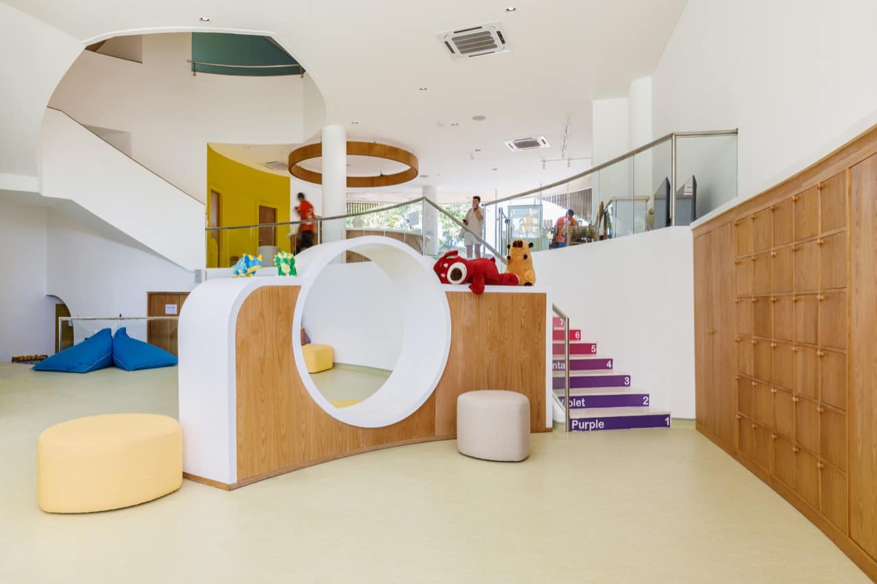 Double-height lobby of the Pangolin Kids Club with a curved white-and-wood reading nook with a circular cut-out, soft poufs, a colour-coded staircase, and a mezzanine railing above