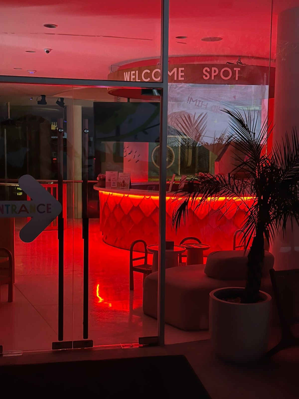 Evening view through the glass entrance doors of the Pangolin Kids Club, with the pangolin-scale facade of the circular reception desk glowing in saturated red RGB light, an entrance arrow on the glass and a potted palm beside lounge poufs