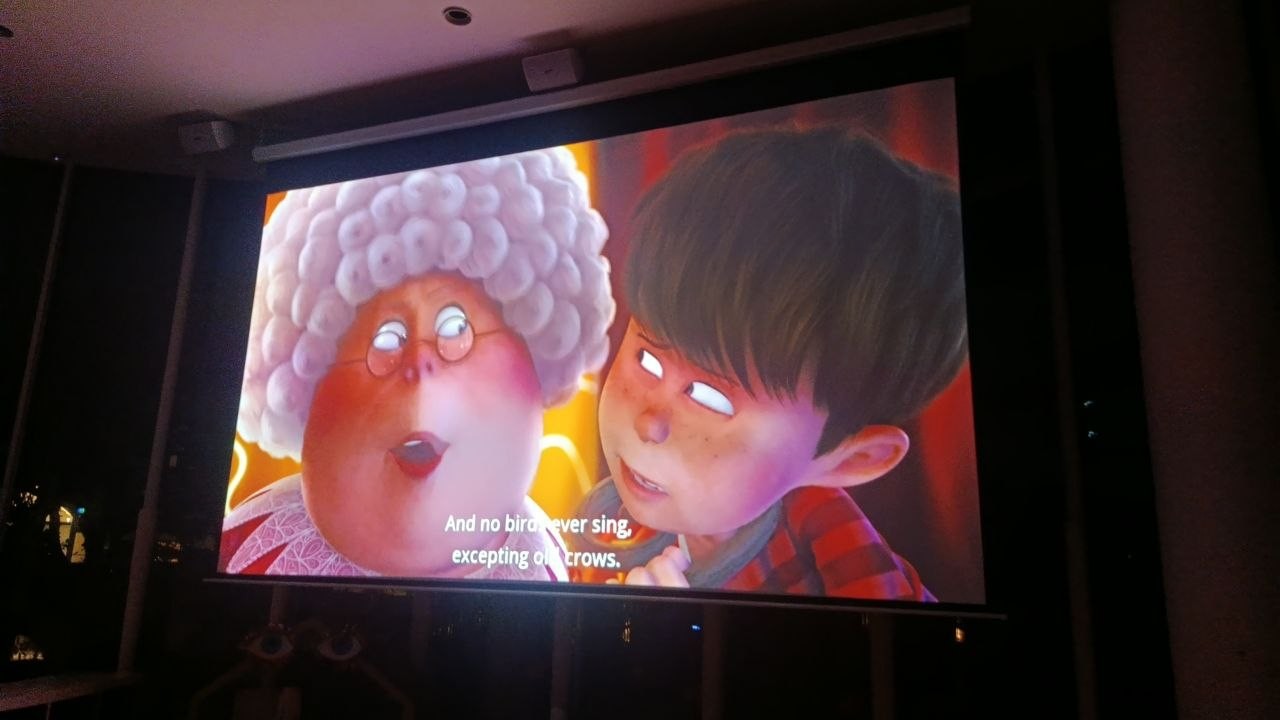 Movie night in the darkened play hall of the Pangolin Kids Club: an animated film with subtitles fills the motorized cinema screen, with the screen housing and two ceiling speakers visible above