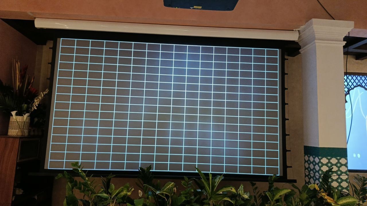 Turquoise calibration test grid held square on the projection screen at SILK Restaurant, Canggu, after 4-corner geometric correction, with the motorized screen housing and the edge of the ceiling projector visible above
