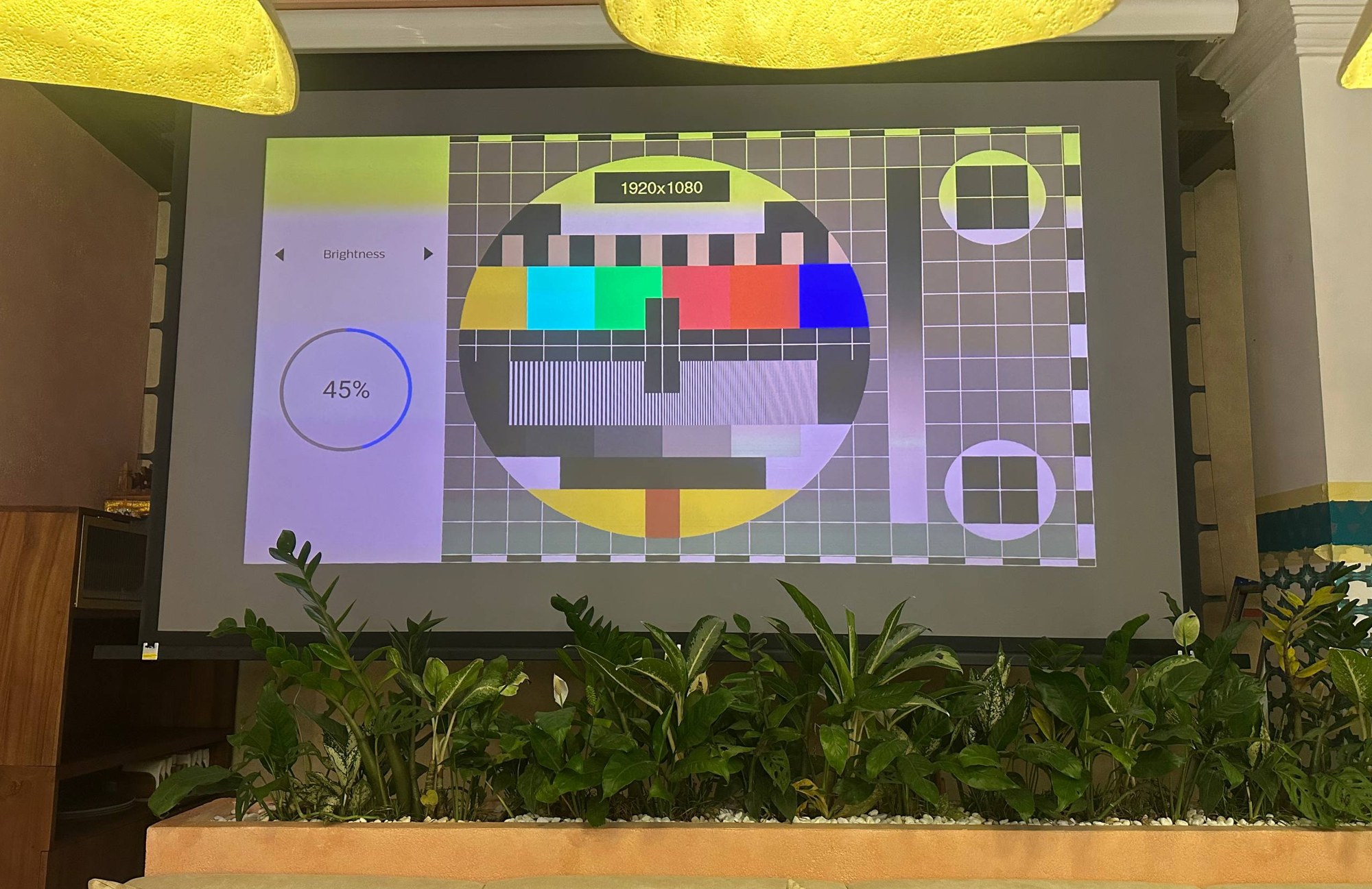 1920×1080 colour-bar test pattern staying legible on the ALR projection screen directly beneath lit yellow fabric chandeliers at SILK Restaurant, with a row of planters and lounge seating below the image